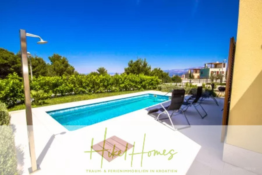 Villa im Mittelmeerstil mit 3 SZ, Pool, Dachterrasse mit Meerblick (2) - Ein moderner Außenpool mit Liegestühlen und einer Außendusche, umgeben von viel Grün und einem klaren blauen Himmel. Im Hintergrund sind moderne Häuser und Berge zu sehen. Der Schriftzug "Hikel Homes" ist am unteren Rand eingeblendet. (45785)