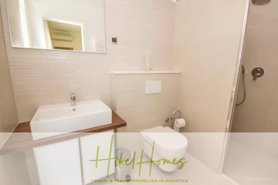 Villa im Mittelmeerstil mit 3 SZ, Pool, Dachterrasse mit Meerblick (2) - Modernes Badezimmer mit weißem Waschbecken, Toilette und begehbarer Dusche; hellbeige Fliesen und eine Arbeitsplatte aus Holz sorgen für einen sauberen, minimalistischen Look. Das "Hikel Homes"-Logo ist am unteren Rand des Bildes eingeblendet. (45801)