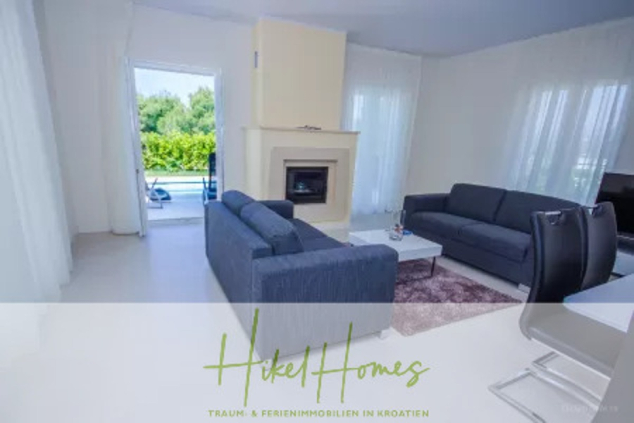 Villa im Mittelmeerstil mit 3 SZ, Pool, Dachterrasse mit Meerblick (2) - Modernes Wohnzimmer mit zwei dunkelgrauen Sofas, einem kleinen Couchtisch auf einem Teppich, einem Kamin, großen Fenstern mit durchsichtigen weißen Vorhängen und Blick auf Grünflächen und einen Pool draußen. Die Textüberlagerung lautet "Hikel Homes". (45795)