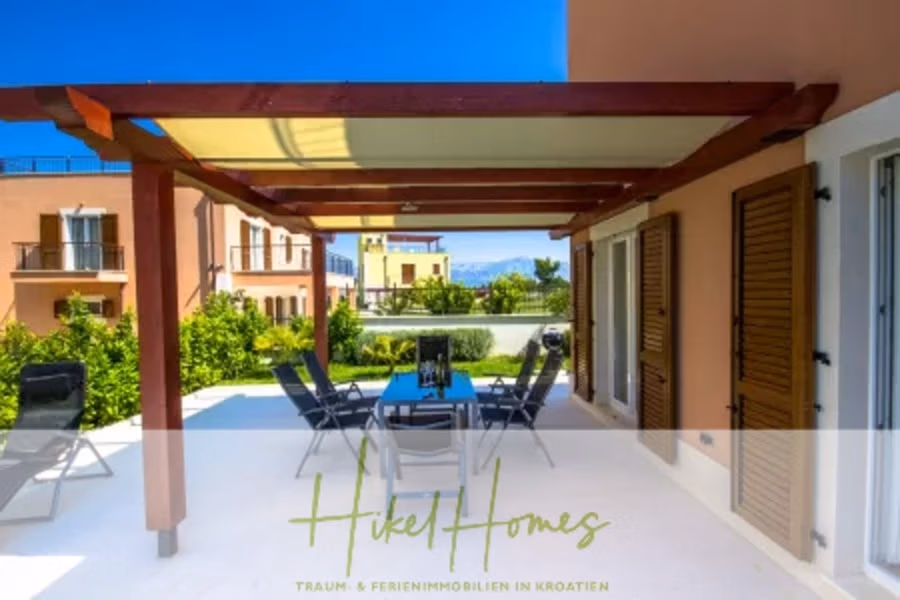 Villa im Mittelmeerstil mit 3 SZ, Pool, Dachterrasse mit Meerblick (1) - Eine moderne Terrasse mit Holzpergola und Gartenmöbeln bietet Blick auf einen üppigen Garten. Sie grenzt an das Haus mit braunen Fensterläden und Türen und bietet einen malerischen Blick auf entfernte Gebäude und einen klaren Himmel. (40949)