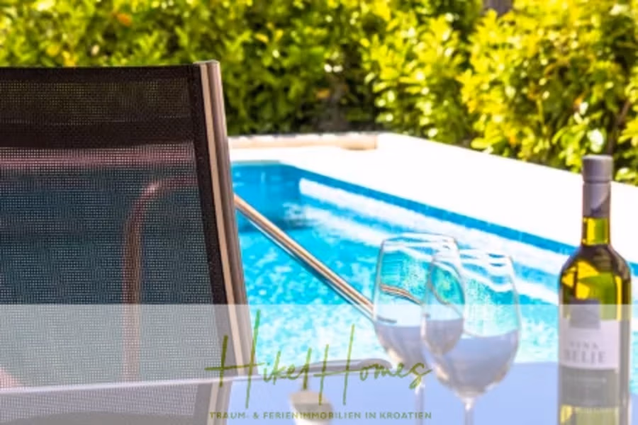 Villa im Mittelmeerstil mit 3 SZ, Pool, Dachterrasse mit Meerblick (1) - Eine sonnenbeschienene Szene im Freien zeigt einen Pool, umgeben von üppigem Grün, eine Flasche Weißwein und zwei Gläser auf einem Tisch. Im Vordergrund ist ein Teil eines Stuhls zu sehen. (40954)