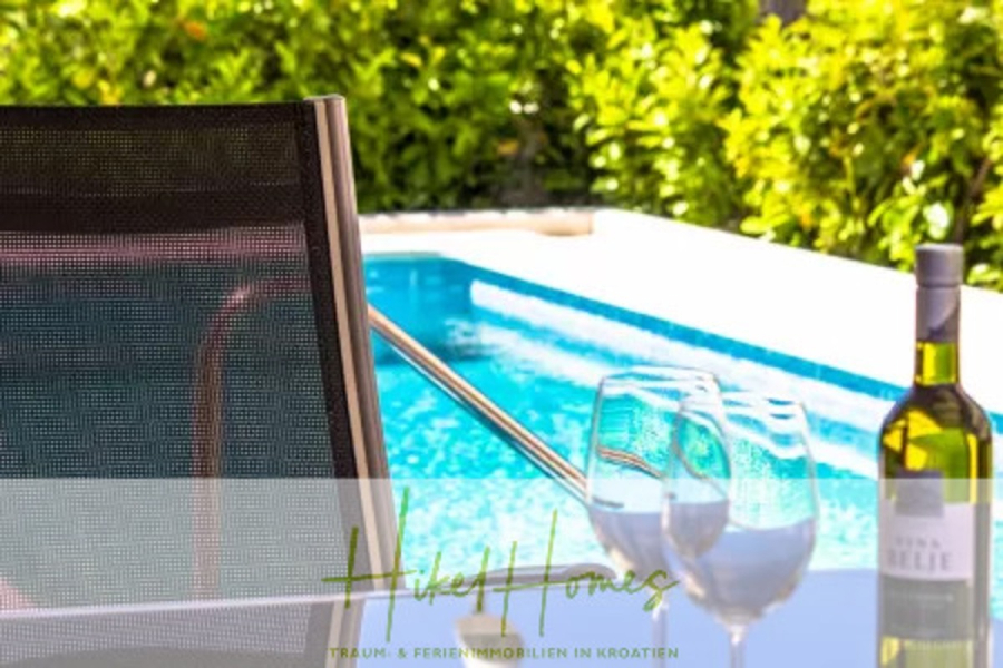 Villa im Mittelmeerstil mit 3 SZ, Pool, Dachterrasse mit Meerblick (1) - Ein moderner Stuhl steht an einem glitzernden Pool, zwei Weingläser und eine Flasche Weißwein stehen auf einem Glastisch, umgeben von üppigem Grün im Hintergrund. (44777)