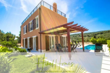 Eine moderne zweistöckige Villa mit großen Fenstern, einer Pergola-überdachten Terrasse, einem Essbereich im Freien und einem Swimmingpool, umgeben von Grün unter einem klaren blauen Himmel. Der Schriftzug "Hikel Homes" erscheint am unteren Rand. - Villa im Mittelmeerstil mit 3 SZ, Pool, Dachterrasse mit Meerblick (1)