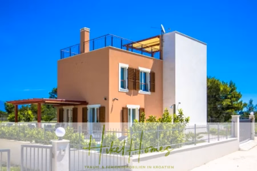 Villa im Mittelmeerstil mit 3 SZ, Pool, Dachterrasse mit Meerblick (1) - Ein zweistöckiges Haus mit Terrakotta- und weißen Außenwänden, braunen Fensterläden und einer Dachterrasse. Es verfügt über einen kleinen Garten, einen Metallzaun und steht vor einem klaren blauen Himmel und Bäumen im Hintergrund. (40951)