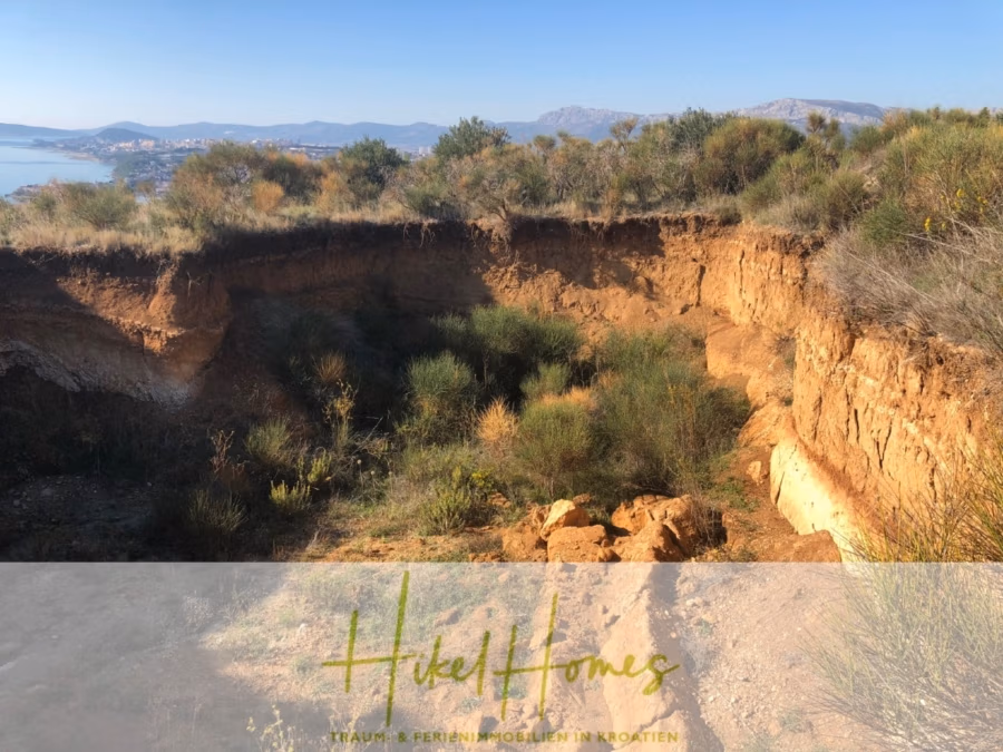 Absolute Alleinlage mit Blick auf Meer, Insel Brac und Split – 511m² auf über 3100m² Grundstück - Ein malerischer Blick auf eine sonnenbeschienene, felsige Landschaft mit spärlicher Vegetation. Im Hintergrund sind Wasser und Berge unter einem klaren, blauen Himmel in der Ferne zu sehen. Im Vordergrund ist eine Grube mit steilen Erdwänden zu sehen. (43247)