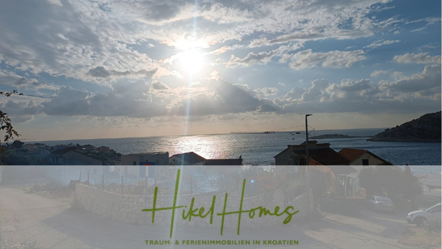 Einziehen im Sommer – Meerblick Neubau-Luxusvilla mit Einrichtung, Infinitypool & Raumkonzept - Eine malerische Küstenansicht bei Sonnenuntergang mit Sonnenlicht, das durch Wolken über dem Meer strömt, Dächern im Vordergrund und dem Text "Hikel Homes Traum- & Ferienimmobilien in Kroatien" über einem transparenten Banner am unteren Rand. (44658)