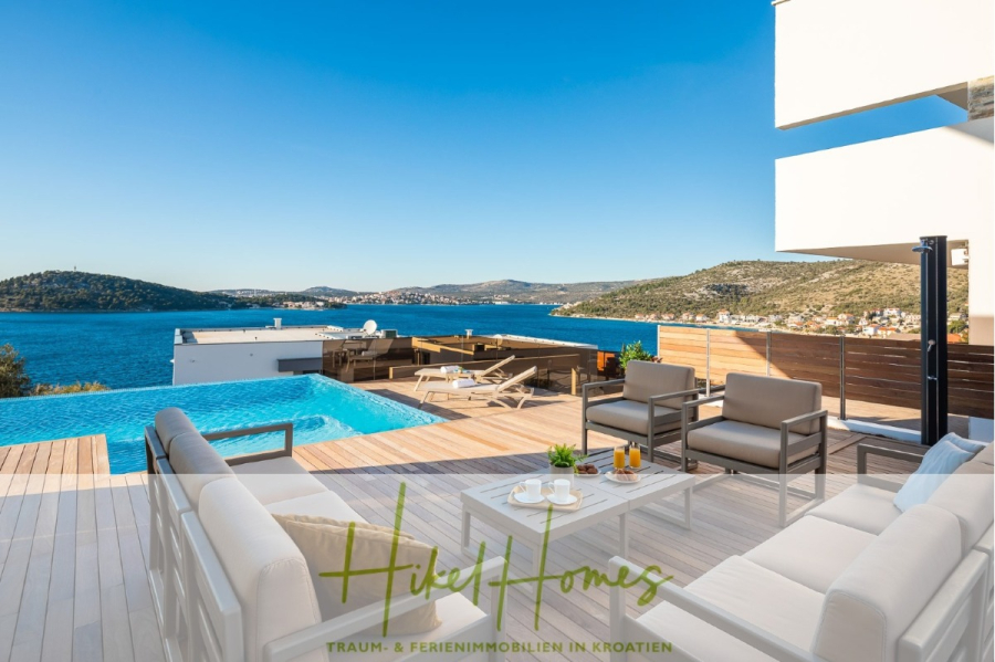 Bezugsfertige Luxus-Meerblick Villa mit 4 SZ, Pool, Sauna, Fitness, Jacuzzi, vollständig möbliert - Moderne Außenterrasse mit Pool mit Blick auf das blaue Meer und die hügelige Küste, mit stilvollen Liegestühlen, Tischen mit Getränken und einem klaren, sonnigen Himmel. Der Text "Hikel Homes" erscheint am unteren Rand. (45624)