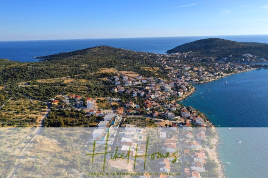 Bezugsfertige Luxus-Meerblick Villa mit 4 SZ, Pool, Sauna, Fitness, Jacuzzi, vollständig möbliert - Luftaufnahme einer Küstenstadt in Kroatien. Zahlreiche Häuser liegen zwischen üppig grünen Hügeln und der Adria. Das Meer glitzert unter einem klaren blauen Himmel, und in Ufernähe sind einige Boote zu sehen. (42253)