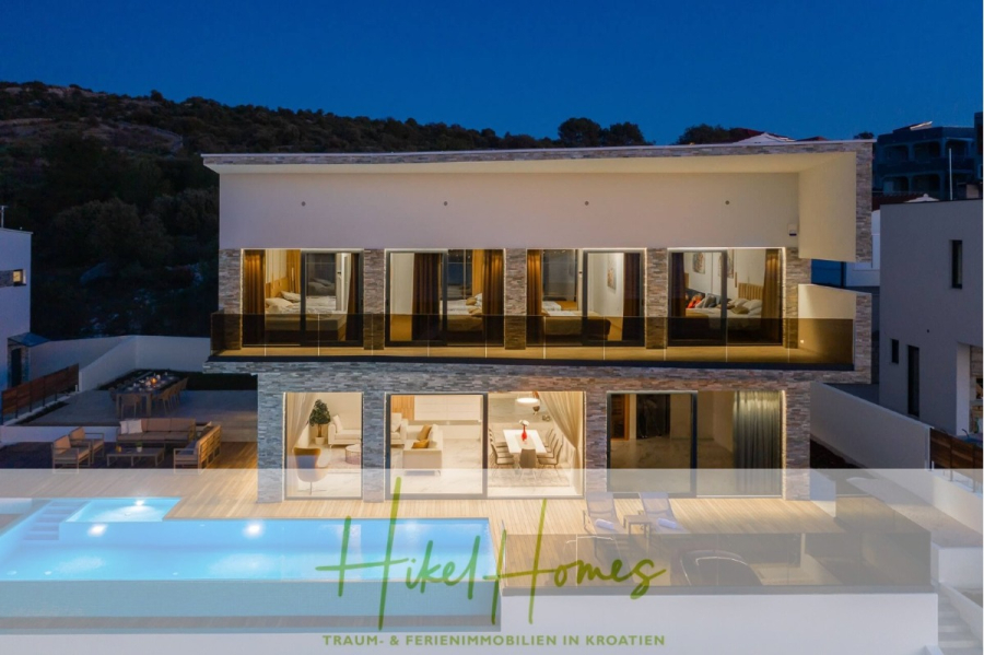 Bezugsfertige Luxus-Meerblick Villa mit 4 SZ, Pool, Sauna, Fitness, Jacuzzi, vollständig möbliert - Moderne zweistöckige Villa mit großen Glasfenstern, die in der Abenddämmerung beleuchtet werden. Das Haus verfügt über geräumige Balkone, einen Außenpool und einen Lounge-Bereich, umgeben von Hügeln. Der Logo-Text am unteren Rand lautet "Hikel Homes: Traum & Ferienimmobilien in Kroatien. (45622)