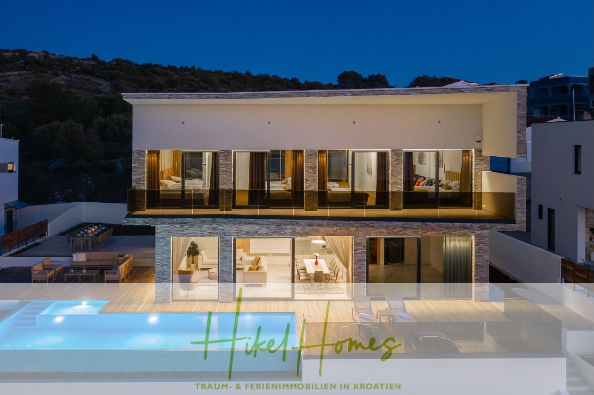 Moderne zweistöckige Villa mit großen Glasfenstern, die in der Abenddämmerung beleuchtet werden. Das Haus verfügt über geräumige Balkone, einen Außenpool und einen Lounge-Bereich, umgeben von Hügeln. Der Logo-Text am unteren Rand lautet "Hikel Homes: Traum & Ferienimmobilien in Kroatien. - Bezugsfertige Luxus-Meerblick Villa mit 4 SZ, Pool, Sauna, Fitness, Jacuzzi, vollständig möbliert
