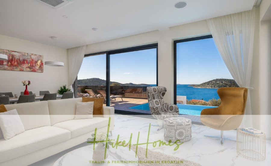 Bezugsfertige Luxus-Meerblick Villa mit 4 SZ, Pool, Sauna, Fitness, Jacuzzi, vollständig möbliert - Modernes Wohnzimmer mit weißem Sofa, goldenem Sessel und Essecke; große Glastüren öffnen sich zu einer Terrasse mit Pool und Liegestühlen, mit Blick auf das Meer und die grünen Hügel unter einem klaren blauen Himmel. (45630)