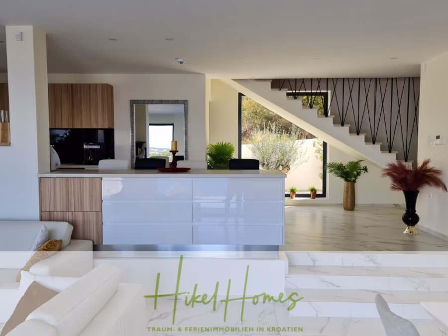 Bezugsfertige Luxus-Meerblick Villa mit 4 SZ, Pool, Sauna, Fitness, Jacuzzi, vollständig möbliert - Moderner, offener Wohnraum mit eleganter Küche, Holzschränken und weißer Kochinsel. Ein lichtdurchfluteter Raum mit Treppe und dekorativen Pflanzen. Unten ist das Logo „Hikel Homes“ mit Text über Immobilien in Kroatien zu sehen. (42258)