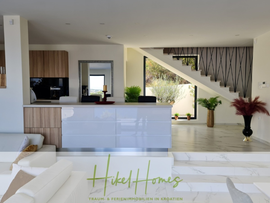 Bezugsfertige Luxus-Meerblick Villa mit 4 SZ, Pool, Sauna, Fitness, Jacuzzi, vollständig möbliert - Moderner offener Küchen- und Wohnbereich mit weißen und hölzernen Schränken, einer verspiegelten Aufkantung, dekorativen Pflanzen und einer Treppe mit schwarzem Geländer. Durch ein großes Fenster unter der Treppe strömt Sonnenlicht herein. (45638)