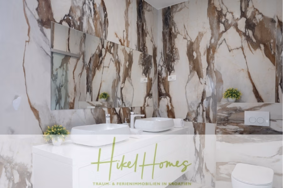 Bezugsfertige Luxus-Meerblick Villa mit 4 SZ, Pool, Sauna, Fitness, Jacuzzi, vollständig möbliert - Luxuriöses Badezimmer mit Marmorwänden, einem weißen Doppelwaschtisch und einem großen Spiegel. Zwei kleine grüne Pflanzen schmücken die Arbeitsplatte. Unten sind die Worte „Hikel Homes“ und weiterer Text eingeblendet. (42266)