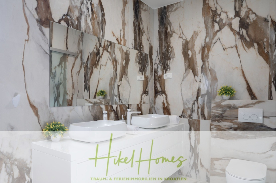 Bezugsfertige Luxus-Meerblick Villa mit 4 SZ, Pool, Sauna, Fitness, Jacuzzi, vollständig möbliert - Modernes Badezimmer mit marmorierten Wänden, zwei weißen Waschbecken mit Spiegeln darüber, kleinen Topfpflanzen auf dem Tresen und dem Regal und einer Toilette auf der rechten Seite. Auf dem Textüberzug steht "Hikel Homes" und "Traum- & Ferienimmobilien in Kroatien". (45647)