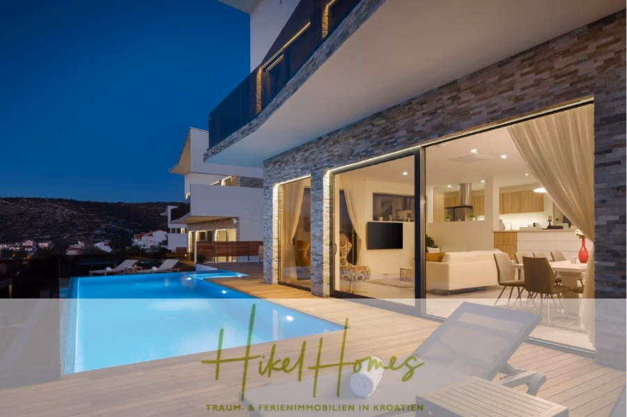 Bezugsfertige Luxus-Meerblick Villa mit 4 SZ, Pool, Sauna, Fitness, Jacuzzi, vollständig möbliert - Moderne Villa mit beleuchtetem Infinity-Pool und großzügiger Terrasse bei Nacht. Der Innenraum ist durch große Glastüren einsehbar und zeigt einen stilvollen Wohnbereich. Im Hintergrund sind Hügel und Gebäude zu sehen. Text unten: „Hikel Homes – Traum- & Ferienimmobilien in Kroatien“. (42260)