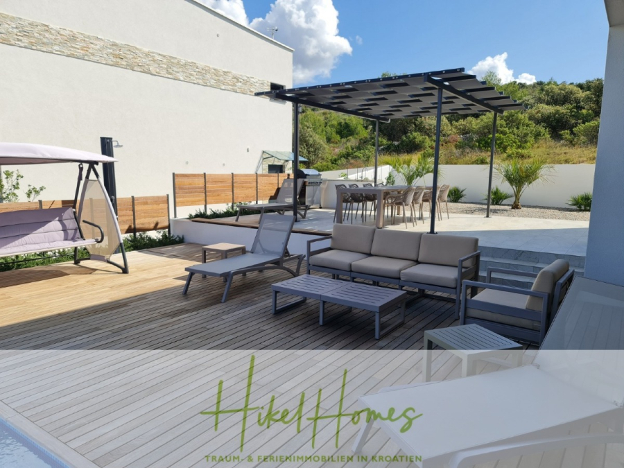 Bezugsfertige Luxus-Meerblick Villa mit 4 SZ, Pool, Sauna, Fitness, Jacuzzi, vollständig möbliert - Moderne Außenterrasse mit Holzbelag, Liegestühlen, einer Schaukel und einem überdachten Essbereich. Grüne Pflanzen säumen die weißen Wände und schaffen eine entspannte Atmosphäre unter einem teilweise bewölkten Himmel. Der Text am unteren Rand lautet "Hikel Homes". (45636)