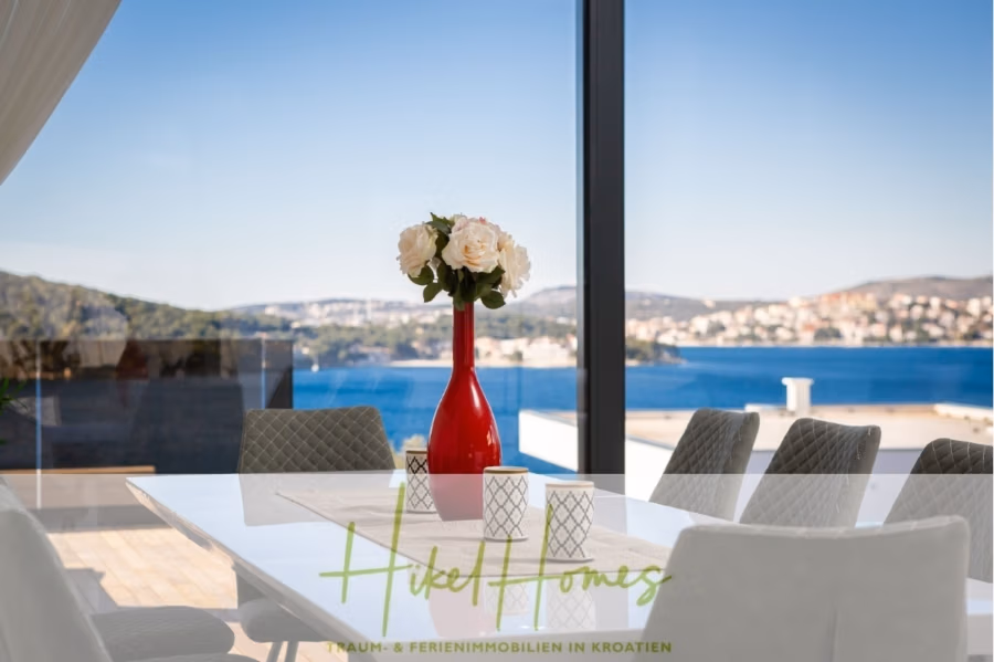 Bezugsfertige Luxus-Meerblick Villa mit 4 SZ, Pool, Sauna, Fitness, Jacuzzi, vollständig möbliert - Ein moderner Essbereich mit weißem Tisch und grauen Stühlen. Eine rote Vase mit weißen Blumen steht auf dem Tisch. Große Fenster bieten einen Panoramablick auf einen malerischen See und Hügel unter einem klaren blauen Himmel. (42270)