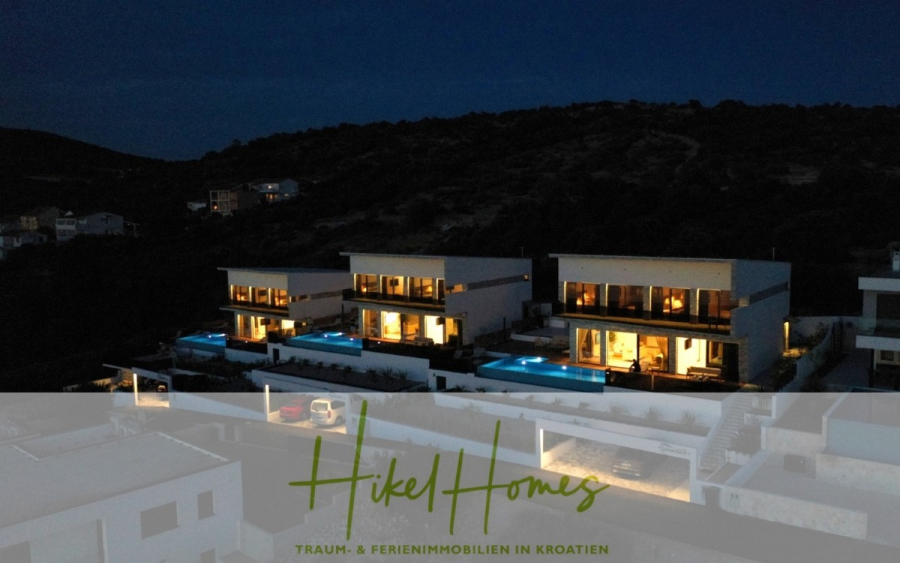 Bezugsfertige Luxus-Meerblick Villa mit 4 SZ, Pool, Sauna, Fitness, Jacuzzi, vollständig möbliert - Ein nächtlicher Blick auf drei moderne Villen mit beleuchteten Pools, die sich an einen Hügel schmiegen. Große Fenster erhellen sie mit warmem Licht. Am unteren Rand des Bildes befindet sich ein halbtransparentes Banner mit dem Text "Hikel Homes". (45655)