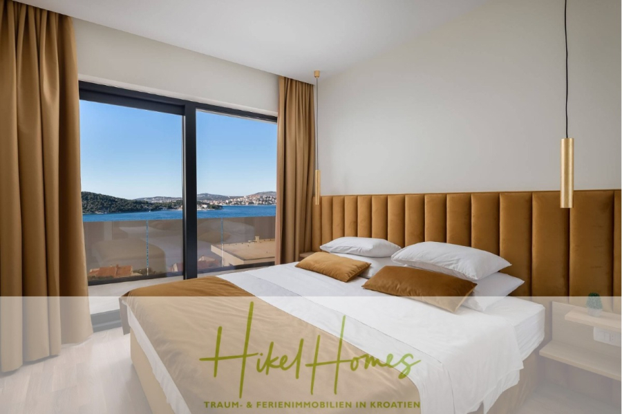 Bezugsfertige Luxus-Meerblick Villa mit 4 SZ, Pool, Sauna, Fitness, Jacuzzi, vollständig möbliert - Ein modernes Schlafzimmer mit einem Doppelbett, goldbraunem Kopfteil und Vorhängen, Pendelleuchten und einem großen Fenster mit Blick auf das Meer und die Küstenstadt. Unten sind "Hikel Homes" und ein deutscher Text eingeblendet. (45644)