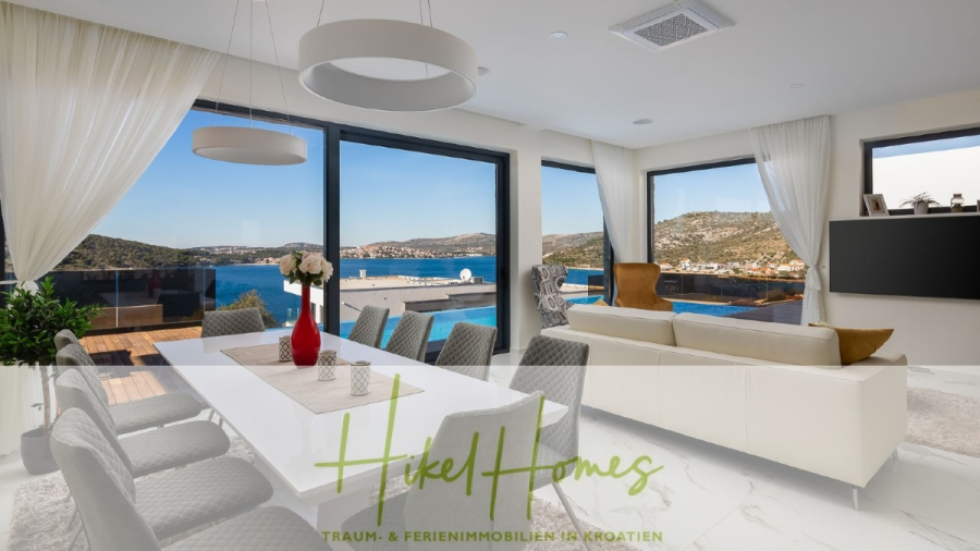 Bezugsfertige Luxus-Meerblick Villa mit 4 SZ, Pool, Sauna, Fitness, Jacuzzi, vollständig möbliert - Moderner, heller Wohn- und Essbereich mit großen Fenstern, die einen atemberaubenden Blick auf das Meer und die Hügel bieten; mit eleganten Möbeln, einem Außenpool und dem Logo von "Hikel Homes" am Boden. (45628)