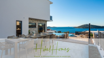 Moderner Essbereich im Freien mit gedecktem Tisch und Blick auf den Pool, die Dächer und das malerische blaue Meer mit grünen Hügeln im Hintergrund; das Branding "Hikel Homes" erscheint am unteren Rand. - Bezugsfertige Luxus-Meerblick Villa mit 4 SZ, Pool, Sauna, Fitness, Jacuzzi, vollständig möbliert