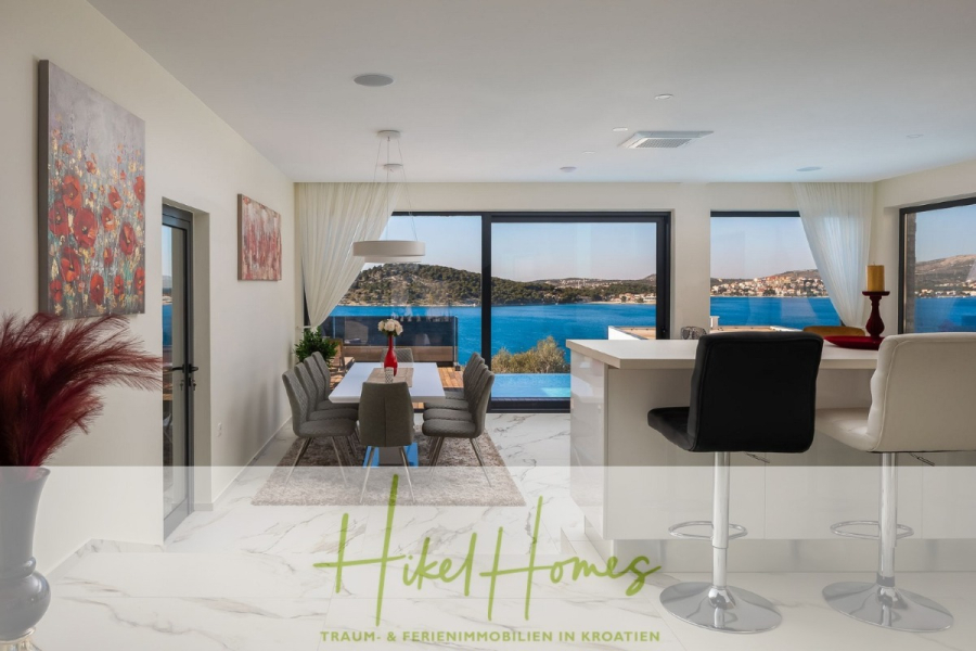 Bezugsfertige Luxus-Meerblick Villa mit 4 SZ, Pool, Sauna, Fitness, Jacuzzi, vollständig möbliert - Moderner offener Küchen- und Essbereich mit großen Fenstern und Blick auf eine blaue Bucht. Weiße Möbel, Barhocker und Wandkunst schaffen einen hellen, eleganten Raum. Das "Hikel Homes"-Logo und der deutsche Text sind am unteren Rand überlagert. (45652)