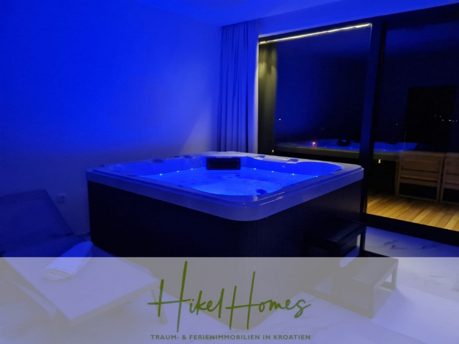 Bezugsfertige Luxus-Meerblick Villa mit 4 SZ, Pool, Sauna, Fitness, Jacuzzi, vollständig möbliert - Ein moderner Innenraum mit einem blau beleuchteten Whirlpool, Liegestühlen und großen Glastüren, die zu einem nächtlichen Balkon führen. Der Text "Hikel Homes Traum- & Ferienimmobilien in Kroatien" erscheint am unteren Rand. (45643)