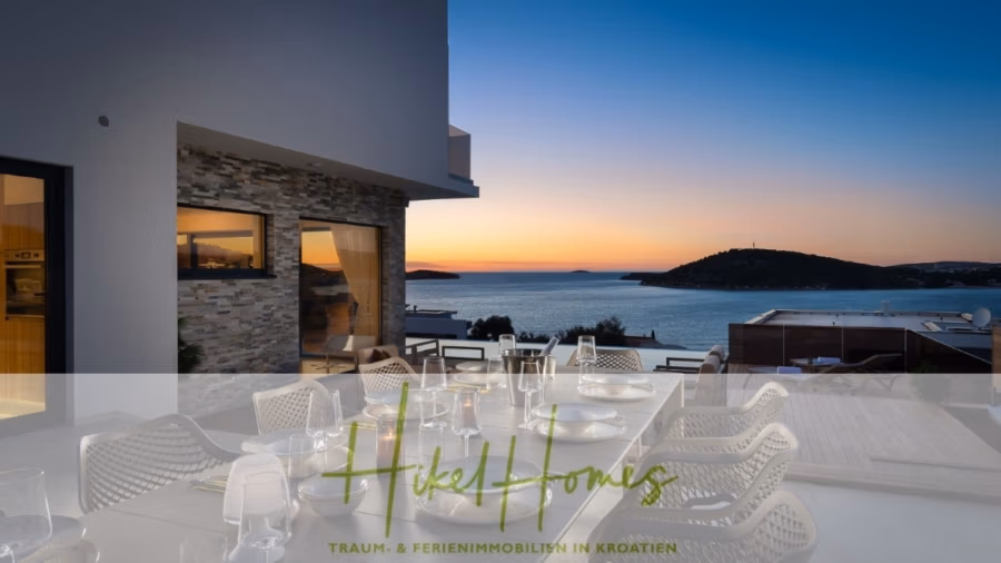 Bezugsfertige Luxus-Meerblick Villa mit 4 SZ, Pool, Sauna, Fitness, Jacuzzi, vollständig möbliert - Essbereich im Freien auf einer Terrasse mit gedecktem Tisch und Blick auf die malerische Küste bei Sonnenuntergang. Links ein modernes Gebäude mit Steinakzenten. Ruhiges Meer und kleine Inseln im Hintergrund unter einem leuchtenden Himmel. (42267)