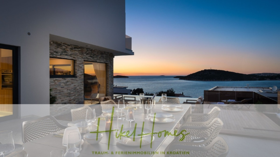 Bezugsfertige Luxus-Meerblick Villa mit 4 SZ, Pool, Sauna, Fitness, Jacuzzi, vollständig möbliert - Ein moderner Essbereich im Freien mit einem gedeckten Tisch bietet einen Blick auf das Meer und die Küstenhügel bei Sonnenuntergang, mit modernen Gebäuden und einem klaren Himmel im Hintergrund. Das Bild ist mit dem Text "Hikel Homes Traum- & Ferienimmobilien in Kroatien" überlagert. (45648)