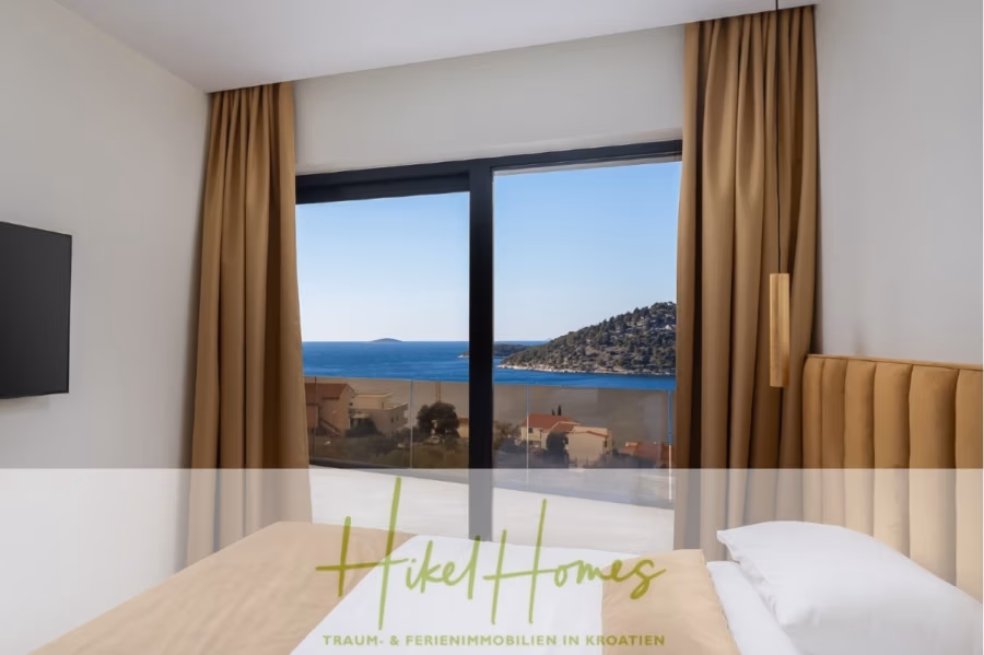 Bezugsfertige Luxus-Meerblick Villa mit 4 SZ, Pool, Sauna, Fitness, Jacuzzi, vollständig möbliert - Blick aus einem modernen Schlafzimmer mit beigen Vorhängen und einem Bett. Durch ein großes Fenster bietet sich ein Panoramablick auf das Meer und die fernen Hügel. Der Text unten lautet: „Hikel Homes“ und erwähnt Ferienimmobilien in Kroatien. (42265)