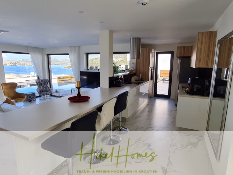 Bezugsfertige Luxus-Meerblick Villa mit 4 SZ, Pool, Sauna, Fitness, Jacuzzi, vollständig möbliert - Moderner offener Küchen- und Wohnbereich mit Meerblick durch große Fenster. Der Raum besticht durch elegante weiße und Holzmöbel, eine lange Kücheninsel mit Barhockern und einen Mix aus modernen Möbeln. Der Raum ist lichtdurchflutet. (42250)