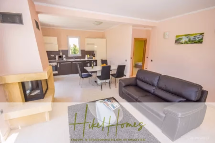 Villa im Mittelmeerstil mit 3 SZ, Pool, Dachterrasse mit Meerblick – möbiliert (15) - Modernes Wohnzimmer und Küche mit rosa Wänden. Zur Ausstattung gehören ein schwarzes Ledersofa, ein Glasesstisch mit acht Stühlen und ein Küchenbereich mit hellen Schränken. Links befindet sich ein Kamin, an der Wand hängt ein Landschaftsfoto. (42337)