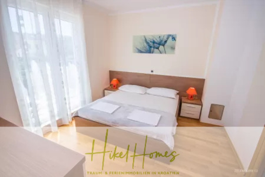 Villa im Mittelmeerstil mit 3 SZ, Pool, Dachterrasse mit Meerblick – möbiliert (15) - Ein helles, modernes Schlafzimmer mit einem Doppelbett, zwei Nachttischen mit roten Lampen, einem großen Fenster mit durchsichtigen Vorhängen, Holzböden und einem Wandkunstwerk über dem Bett. Der Text darüber lautet "Hikel Homes". (45701)