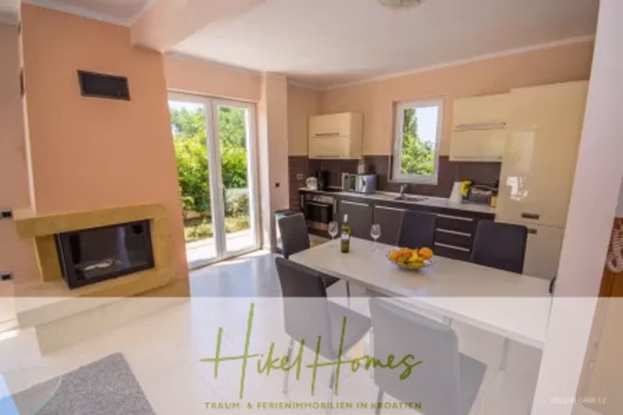Villa im Mittelmeerstil mit 3 SZ, Pool, Dachterrasse mit Meerblick – möbiliert (15) - Ein moderner Küchen- und Essbereich mit großen Fenstern. Der Raum verfügt über helle Wände, einen Kamin auf der linken Seite, einen Tisch mit Stühlen und eine Küche mit Schränken und Geräten. Durch die Fenster blickt man ins Grüne. (42338)