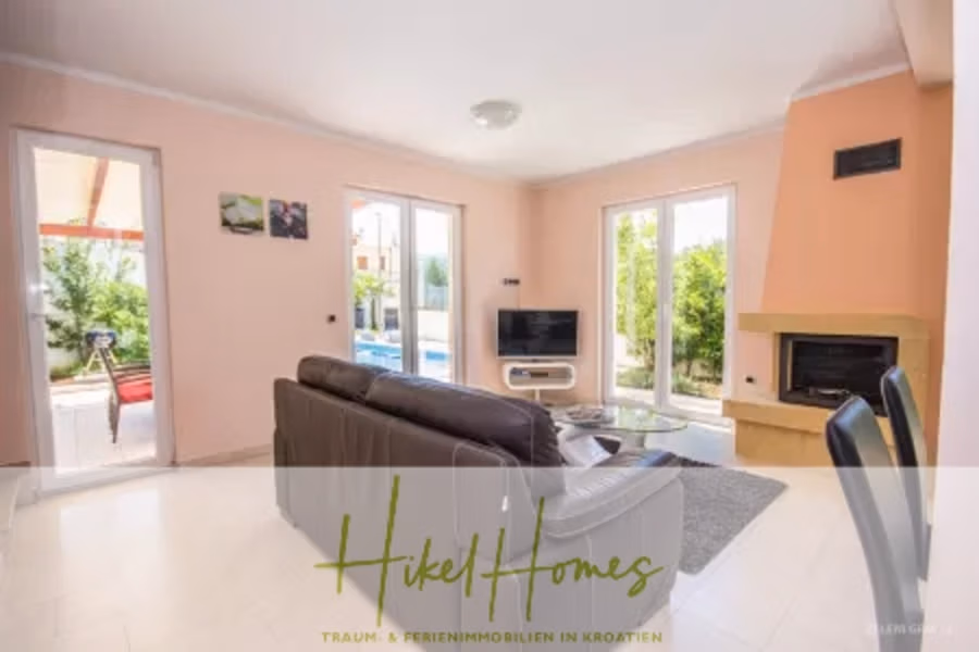 Villa im Mittelmeerstil mit 3 SZ, Pool, Dachterrasse mit Meerblick – möbiliert (15) - Modernes Wohnzimmer mit einem beigen Sofa vor einem an der Wand montierten Fernseher. Große Fenster bieten Ausblick ins Grüne. Rechts befindet sich ein Kamin, und ein kleiner Essbereich ist sichtbar. Unten steht „HikelHomes“, darunter deutscher Text. (42333)