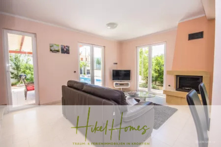 Villa im Mittelmeerstil mit 3 SZ, Pool, Dachterrasse mit Meerblick – möbiliert (15) - Helles Wohnzimmer mit großen Fenstern, einem braunen Ledersofa vor einem Fernseher, einem Kamin, einem schwarzen Stuhl und Blick auf einen Garten und einen Pool. Der Schriftzug "Hikel Homes" ist am unteren Rand eingeblendet. (45694)