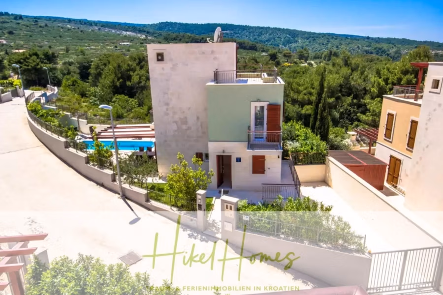 Villa im Mittelmeerstil mit 3 SZ, Pool, Dachterrasse mit Meerblick – möbiliert (15) - Eine kleine, moderne Villa mit Pool, umgeben von viel Grün und einer Waldlandschaft. Die Straße führt um das Grundstück herum, der Himmel ist klar und blau. Die Fassade der Villa ist hell mit einer grünen Akzentwand. (42331)