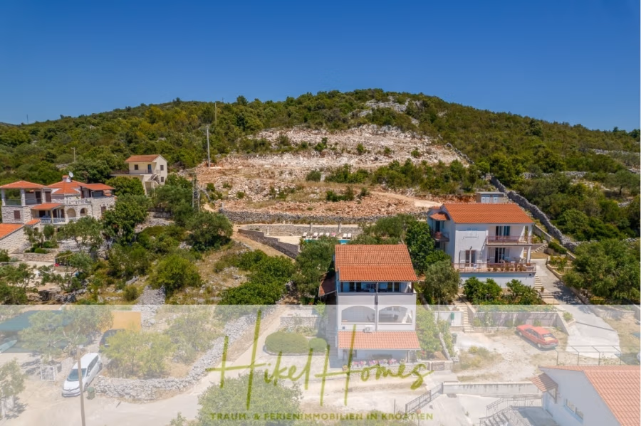 Meerblick Appartementhaus mit 3 Wohnungen, Karten, Terrassen - Erhöhte Ansicht von Häusern, umgeben von üppigem Grün und felsigen Hügeln unter einem klaren blauen Himmel. Am unteren Bildrand ist ein Schild mit der Aufschrift „Fikel Homes“ zu sehen. (40997)