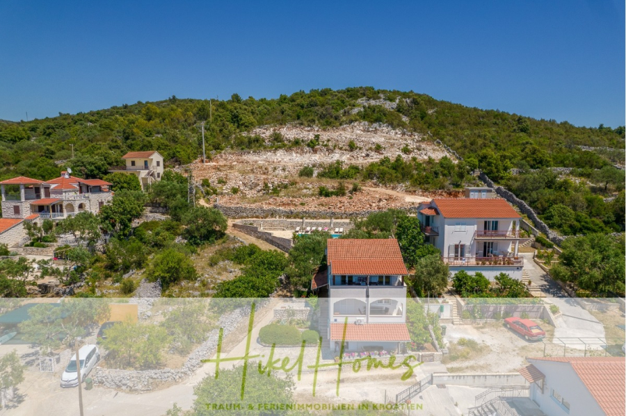 Meerblick Appartementhaus mit 3 Wohnungen, Karten, Terrassen - Luftaufnahme von Häusern, umgeben von Grün und felsigen Hügeln unter einem klaren blauen Himmel, mit dem Logo "Hikel Homes" und einem deutschen Text über Traumferienimmobilien, der unten eingeblendet ist. (45325)
