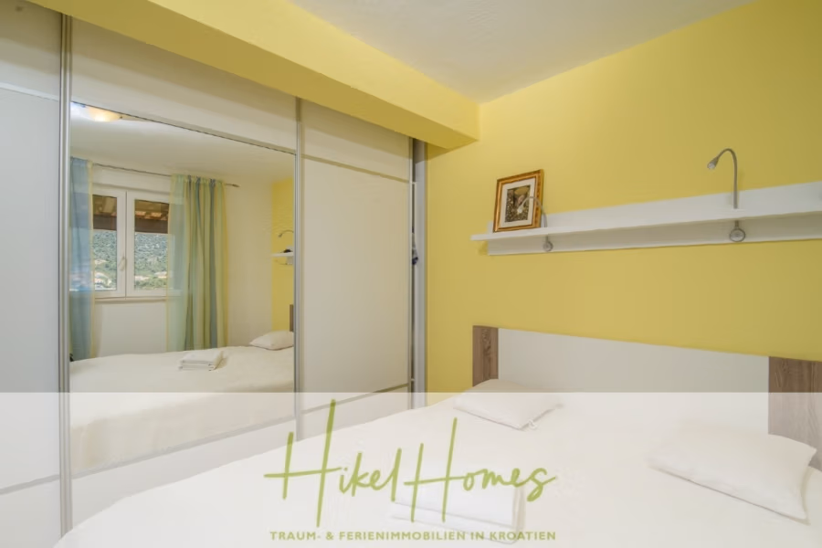 Meerblick Appartementhaus mit 3 Wohnungen, Karten, Terrassen - Ein gemütliches Schlafzimmer mit einer gelben Akzentwand und einem großen Bett mit weißer Bettwäsche. Über dem Bett befindet sich ein Regal mit einem gerahmten Bild. Ein verspiegelter Kleiderschrank spiegelt das Fenster mit malerischer Aussicht wider. Der Text unten lautet: „Hikel Homes – Traum- & Ferienimmobilien in Kroatien.“. (40991)