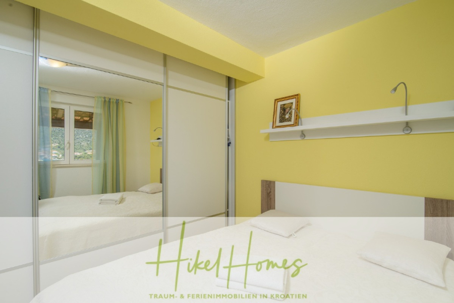 Meerblick Appartementhaus mit 3 Wohnungen, Karten, Terrassen - Helles Schlafzimmer mit einer gelben Akzentwand, einem Doppelbett, einem weißen Regal mit einem Bilderrahmen und einer Lampe, großen verspiegelten Schranktüren und einem Fenster mit grünen und weißen Vorhängen. Das Logo von "Hikel Homes" ist am unteren Rand eingeblendet. (45317)
