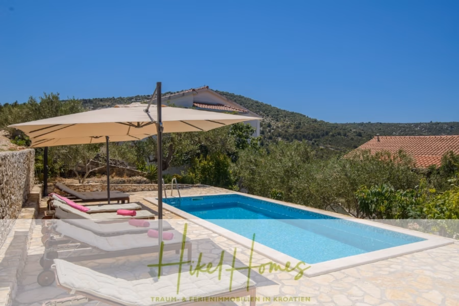 Meerblick Appartementhaus mit 3 Wohnungen, Karten, Terrassen - Blick auf einen mediterranen Garten mit Pool, umgeben von einer Steinterrasse und mehreren weißen Liegestühlen mit rosa Handtüchern. Ein Sonnenschirm spendet Schatten. Im Hintergrund sind Hügel und ein Haus unter einem klaren blauen Himmel zu sehen. (40987)