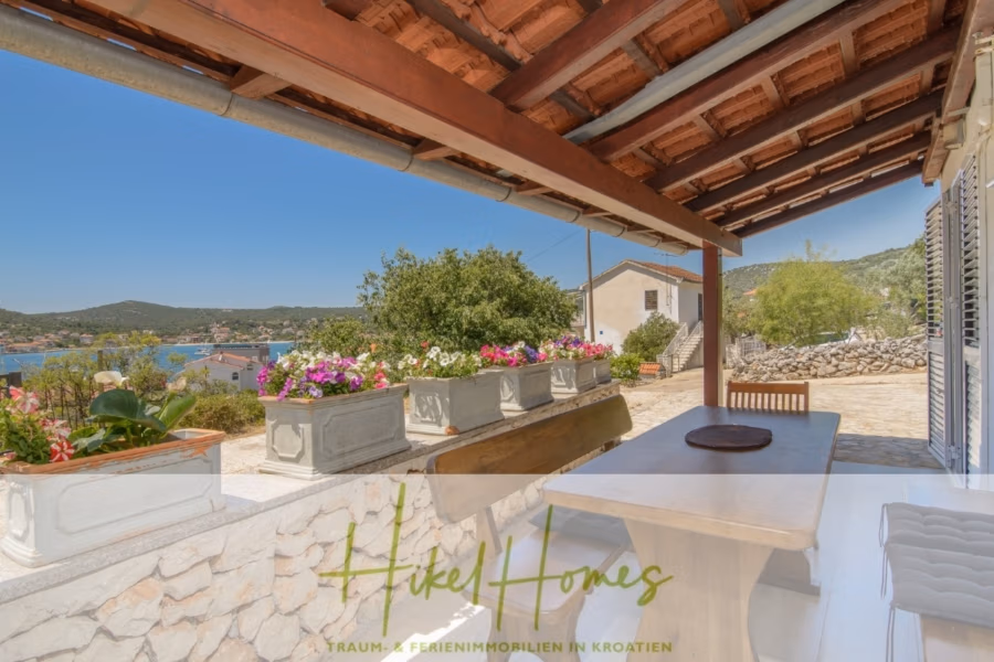 Meerblick Appartementhaus mit 3 Wohnungen, Karten, Terrassen - Überdachte Außenterrasse mit Holztisch und Bänken bietet einen malerischen Blick auf Meer und Hügel. Blumenkästen mit bunten Blüten schmücken das Geländer. Steinmauern und ein Ziegeldach, im Hintergrund ist ein weiteres Gebäude zu sehen. (40994)