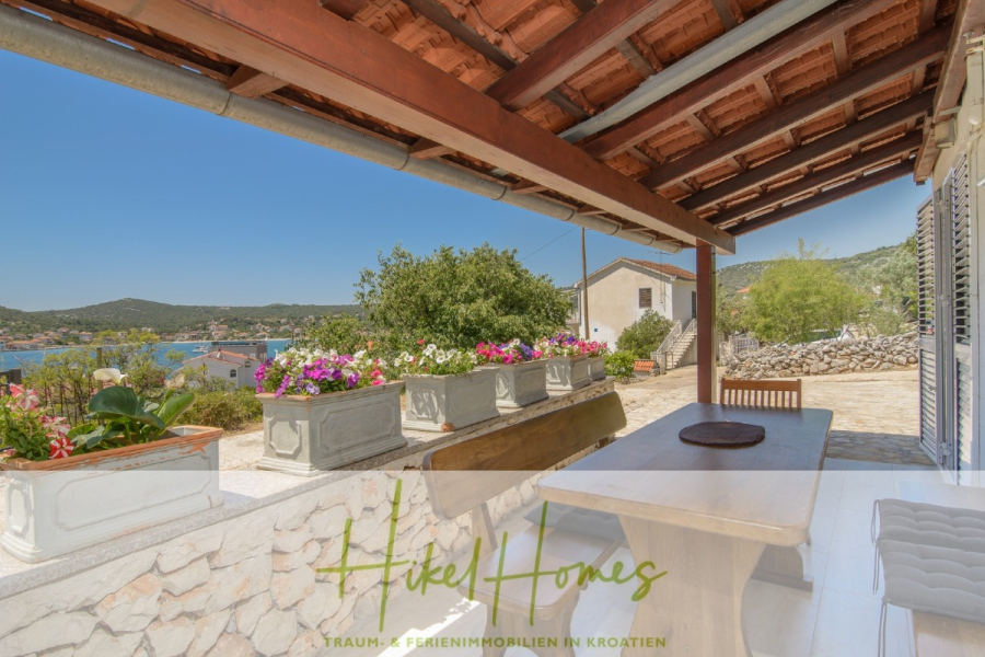 Meerblick Appartementhaus mit 3 Wohnungen, Karten, Terrassen - Eine überdachte Außenterrasse mit Holztisch und Bänken bietet einen Blick auf die malerischen Hügel und das Wasser. Blumenkästen säumen das steinerne Geländer, und unter dem sonnigen Himmel sind die traditionellen Ziegeldächer zu sehen. (45321)