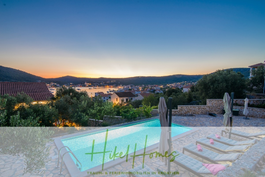 Meerblick Appartementhaus mit 3 Wohnungen, Karten, Terrassen - Infinity-Pool mit Liegestühlen mit Blick auf ein malerisches Küstendorf bei Sonnenuntergang, umgeben von Hügeln und Grünflächen, mit ruhigem Wasser und sanftem Abendlicht. Das Logo von "Hikel Homes" und der deutsche Text erscheinen am unteren Rand des Bildes. (45322)