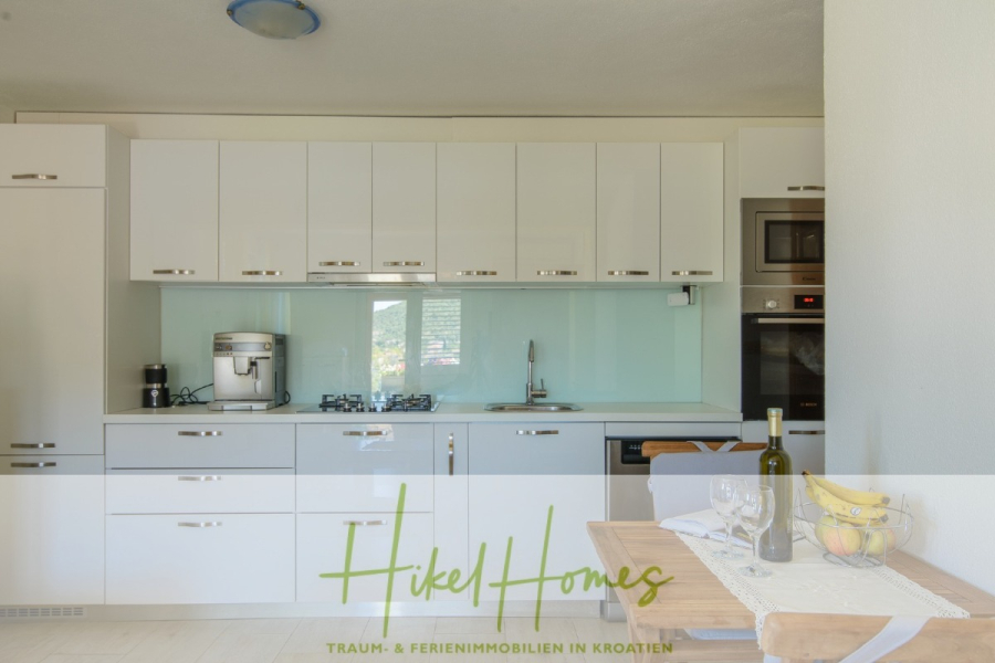 Meerblick Appartementhaus mit 3 Wohnungen, Karten, Terrassen - Moderne weiße Küche mit Glasaufkantung, Einbaubackofen, Gasherd und Arbeitsplattengeräten. Im Vordergrund ist ein kleiner Esstisch mit Wein, Bananen und Geschirr gedeckt. Das Sonnenlicht erhellt den Raum. (45318)