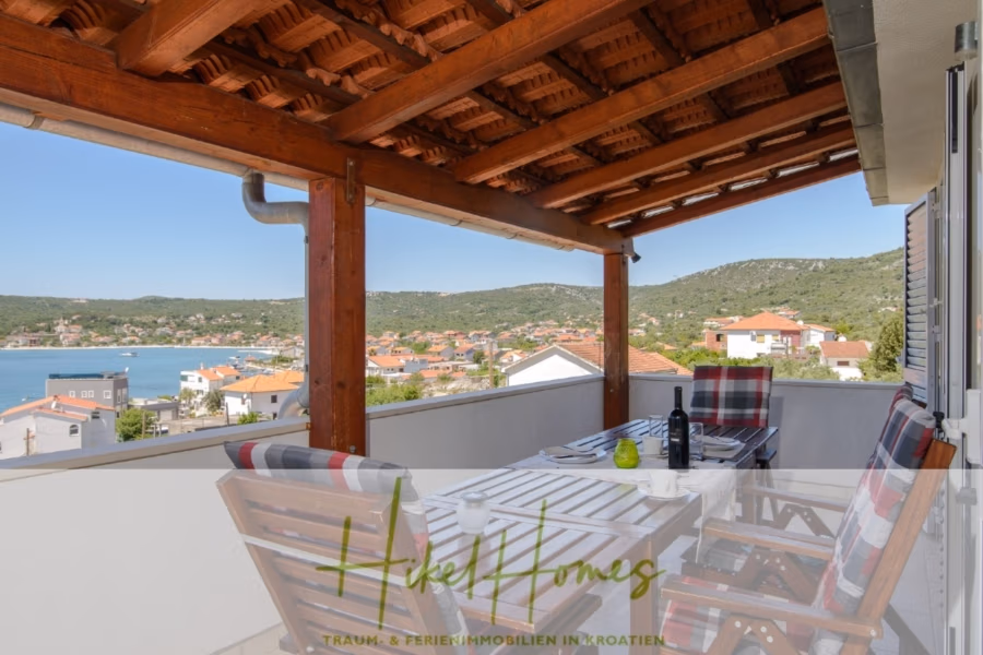 Meerblick Appartementhaus mit 3 Wohnungen, Karten, Terrassen - Überdachter Balkon mit Holzbalken und Blick auf eine malerische Bucht und die Häuser am Hang. Ein Tisch mit weißer Tischdecke, Wein und karierten Kissen auf Holzstühlen bietet eine gemütliche Atmosphäre für Mahlzeiten im Freien. Der strahlend blaue Himmel trägt zur ruhigen Atmosphäre bei. (40980)