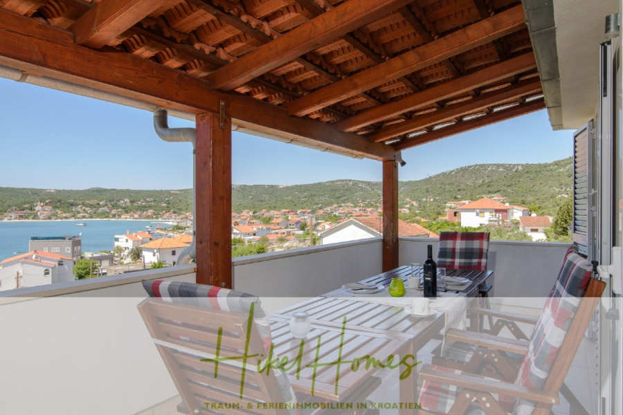 Meerblick Appartementhaus mit 3 Wohnungen, Karten, Terrassen - Überdachter Balkon mit Holzdach, Tisch für vier Personen, gepolsterten Stühlen und einer malerischen Aussicht auf eine Küstenstadt und Hügel unter einem klaren Himmel. Das Logo von "Hieke Homes" ist am unteren Rand des Bildes eingeblendet. (45305)