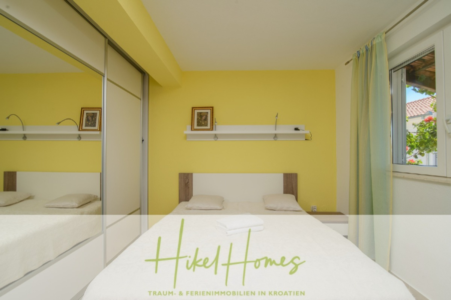 Meerblick Appartementhaus mit 3 Wohnungen, Karten, Terrassen - Ein helles Schlafzimmer mit einem Doppelbett, einer gelben Akzentwand, verspiegelten Schranktüren, zwei gerahmten Bildern, einem Regal über dem Bett mit Lampen, weißem Bettzeug und einem Fenster mit hellgrünen Vorhängen. Der Text am unteren Rand lautet "Hikel Homes". (45314)